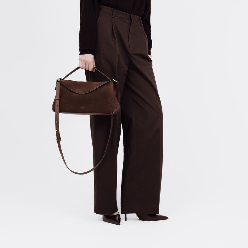 Model holding STOW London's East West bag in Chocolate Suede leather by the top handle at her side featuring gold-coated stainless steel hardware and its detachable crossbody strap on a white background