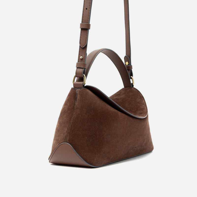 Off-axis view of STOW London's East West in Chocolate suede leather featuring gold-coated stainless steel hardware and its detachable crossbody strap on a light grey background