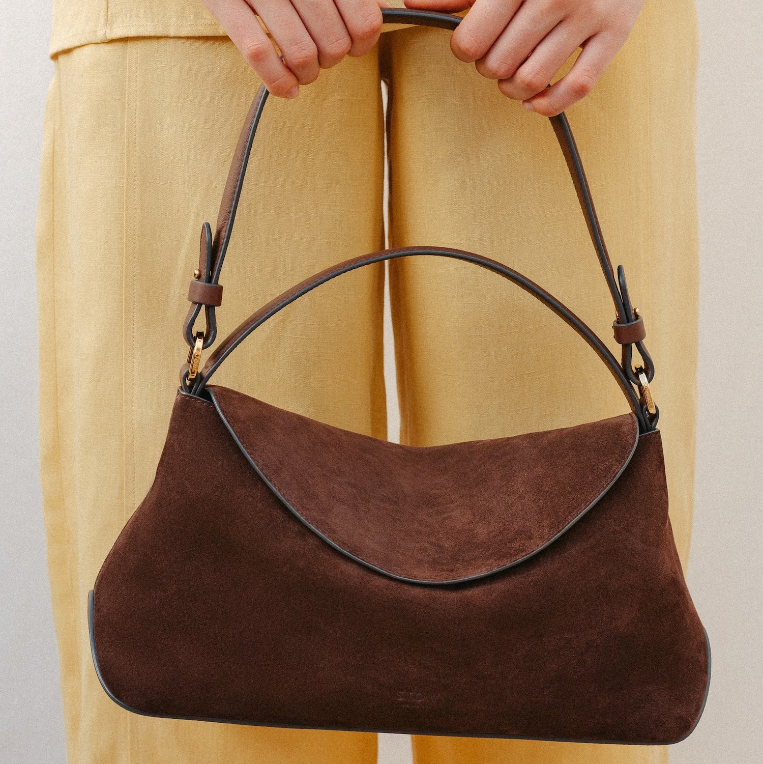 Model holding STOW London's East West in Chocolate suede leather featuring the detachable shoulder strap with gold hardware