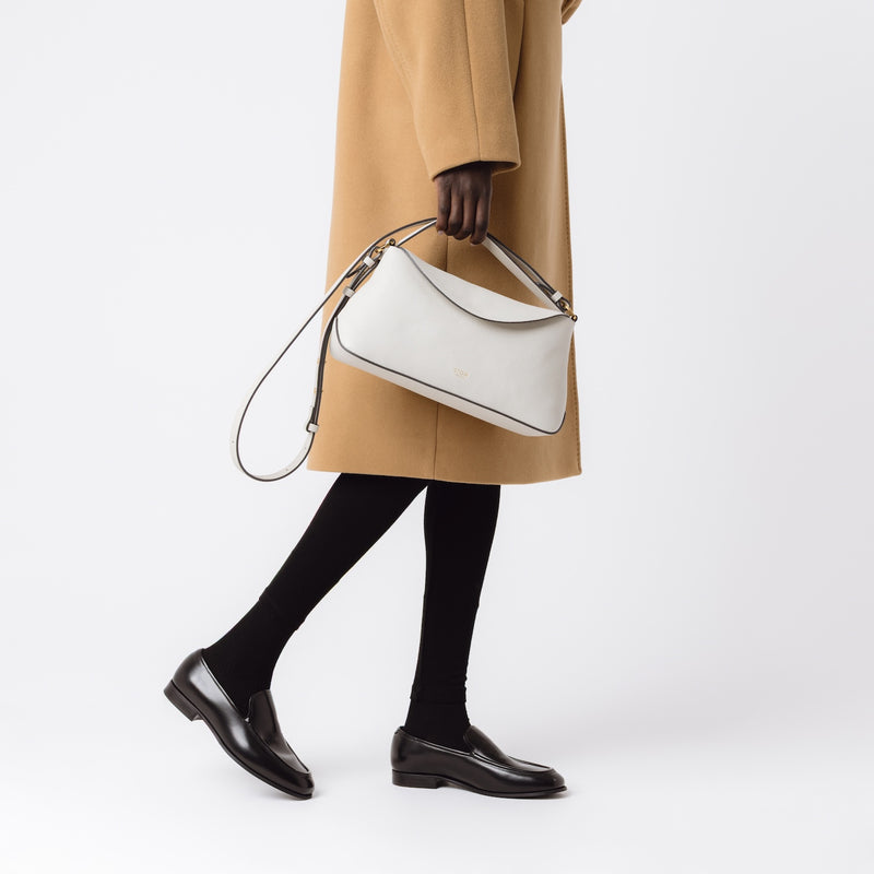 Model holding STOW London's East West in Cloud Grey pebbled leather at her side featuring gold-coated stainless steel hardware on a light grey background