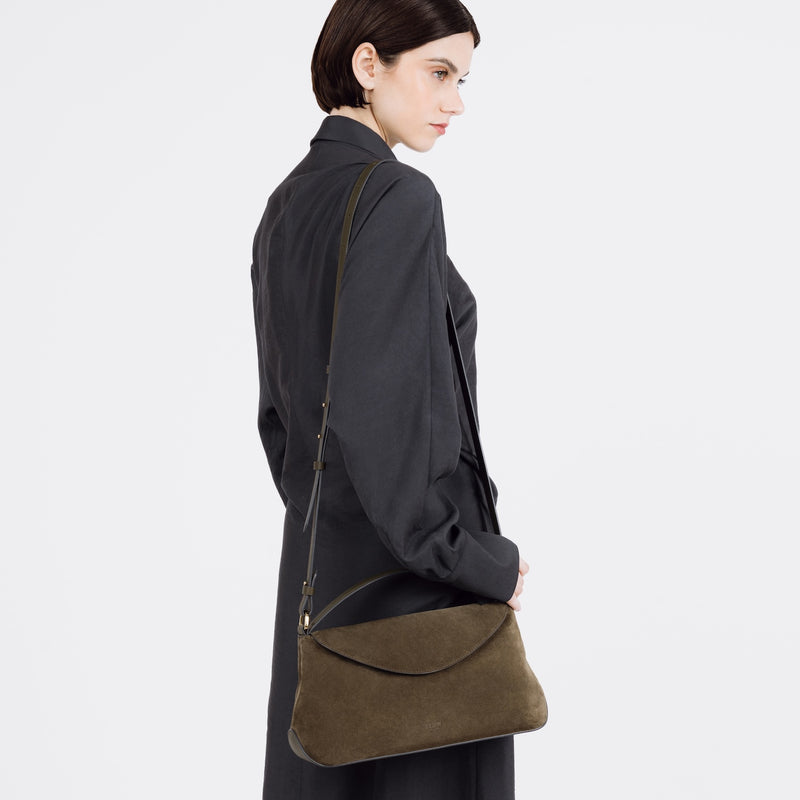 Model wearing STOW London's East West bag in Dark Green Suede leather on her shoulder featuring gold-coated stainless steel hardware on a white background
