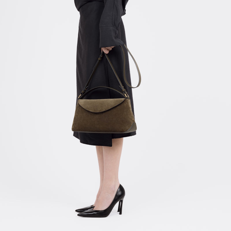 Model holding STOW London's East West bag in Dark Green Suede leather by its detachable, adjustable crossbody strap at her side featuring gold-coated stainless steel hardware on a white background