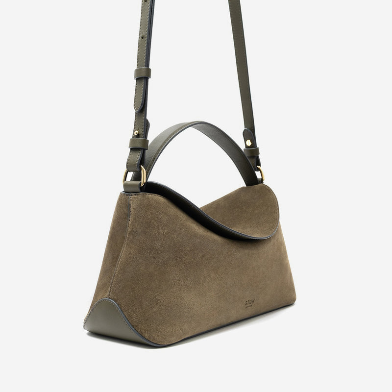 Off-axis view of STOW London's East West in Dark Green suede leather featuring gold-coated stainless steel hardware and detachable crossbody strap on a light grey background