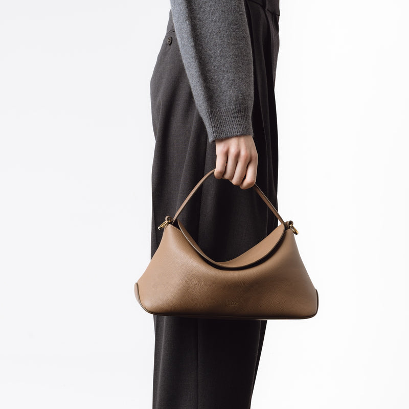 Model holding STOW London's East West in Mocha leather by top handle at her side featuring gold-coated stainless steel hardware on a white background