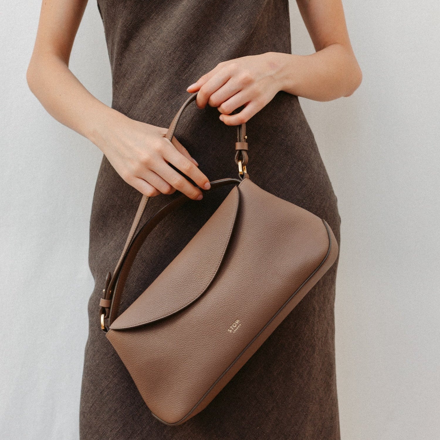Model holding STOW London's East West in Mocha leather featuring the detachable shoulder strap and top handle with gold hardware and details 