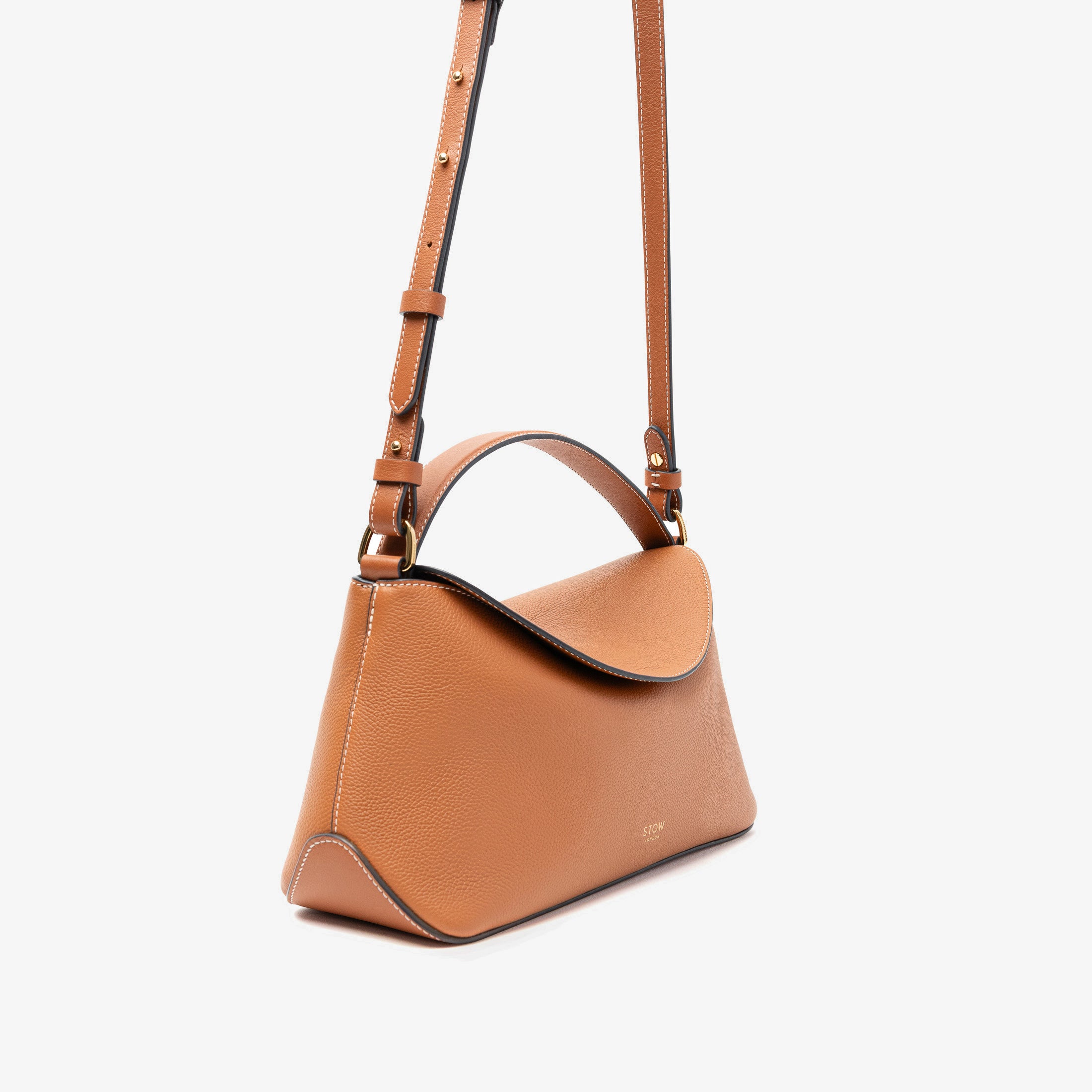 Off-axis view of STOW London's East West bag in Earth Tan pebbled leather and detachable crossbody strap featuring gold-coated stainless steel hardware on a light grey background