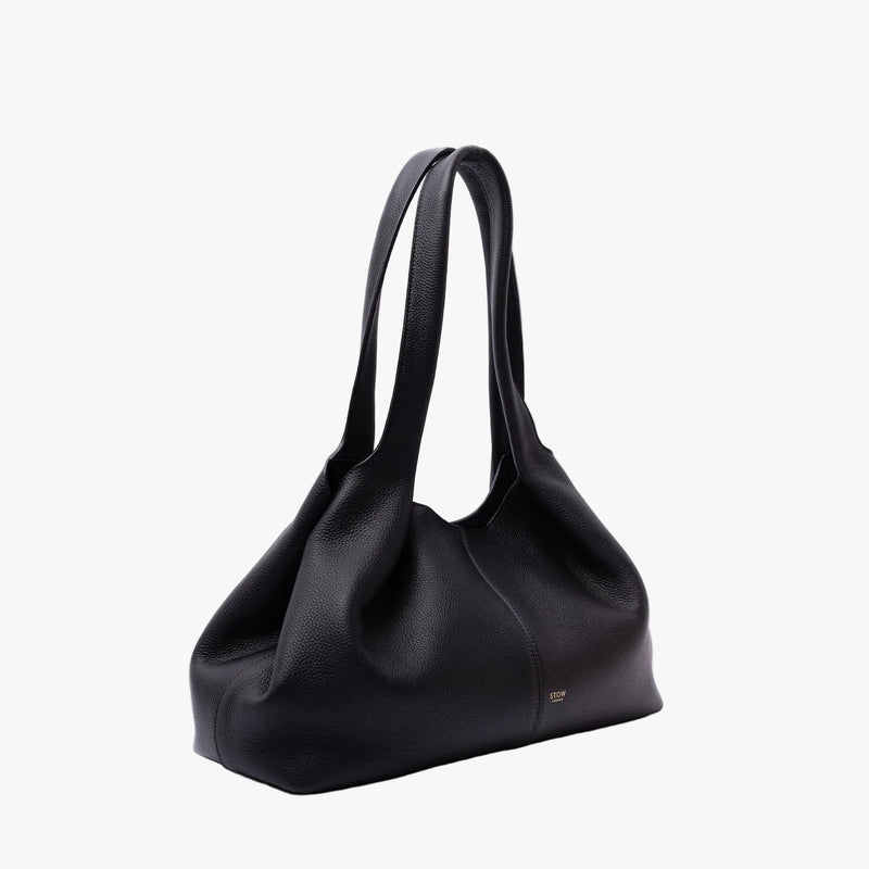 Off-axis view of STOW London's Hobo Tote in Black pebbled leather in a triangular hobo shape on a light grey background featuring a pleated front panel and small gold embossed logo