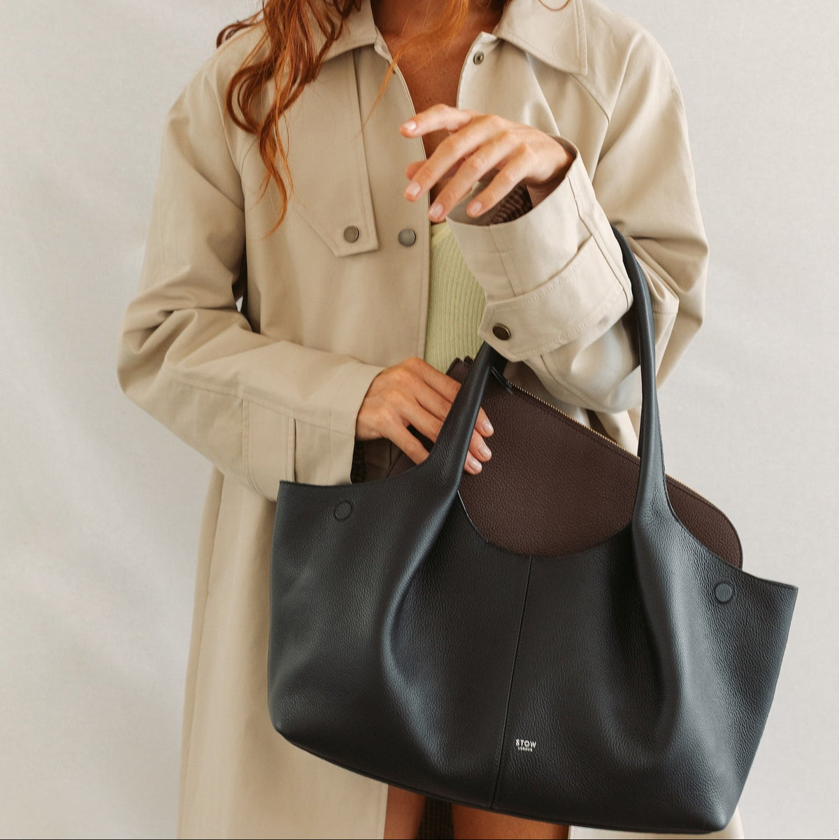 Model with STOW London's Hobo Tote in Black pebbled leather inserting the Midi Leather Sleeve in Espresso into the bag. Featuring a pleated front panel and a silver embossed logo