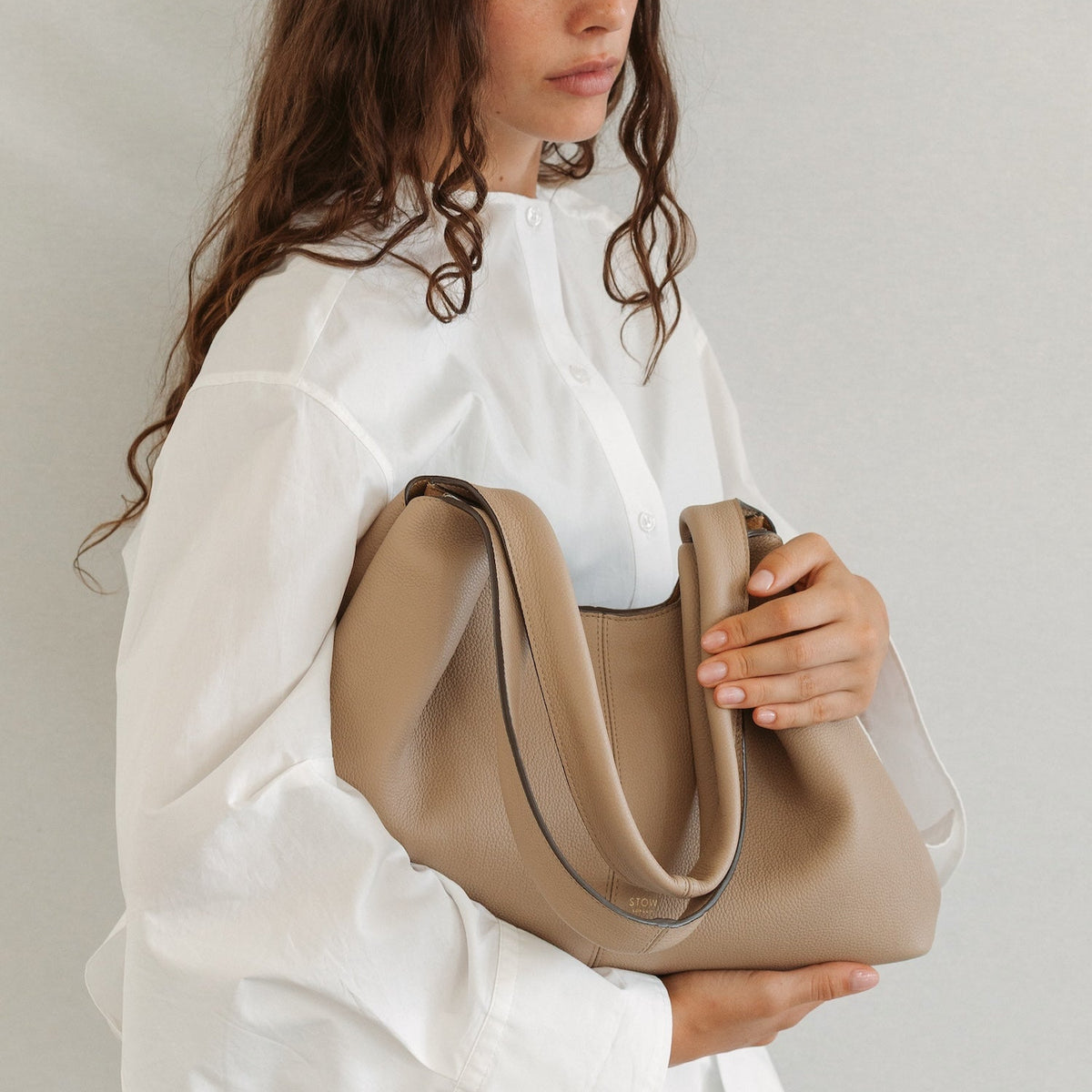 Model holding STOW London's Hobo Tote in Camel pebbled leather featuring a pleated front panel and gold-coated stainless steel hardware 