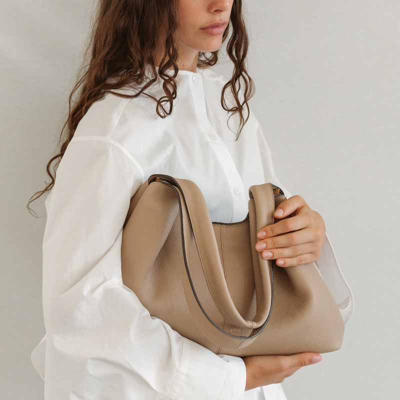 Model holding STOW London's Hobo Tote in Camel pebbled leather featuring a pleated front panel and gold-coated stainless steel hardware 