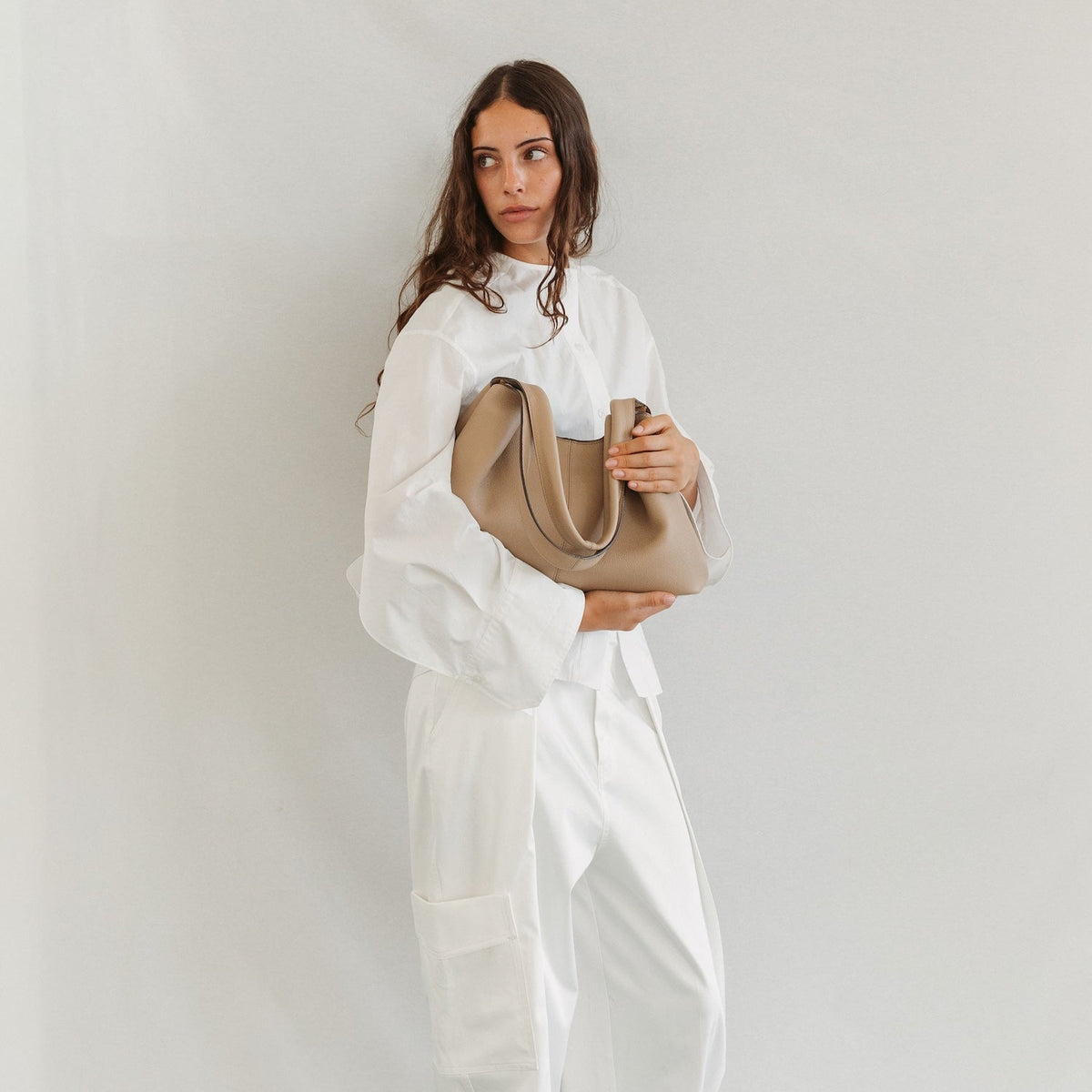 Model holding STOW London's Hobo Tote in Camel pebbled leather featuring a pleated front panel and gold-coated stainless steel hardware 
