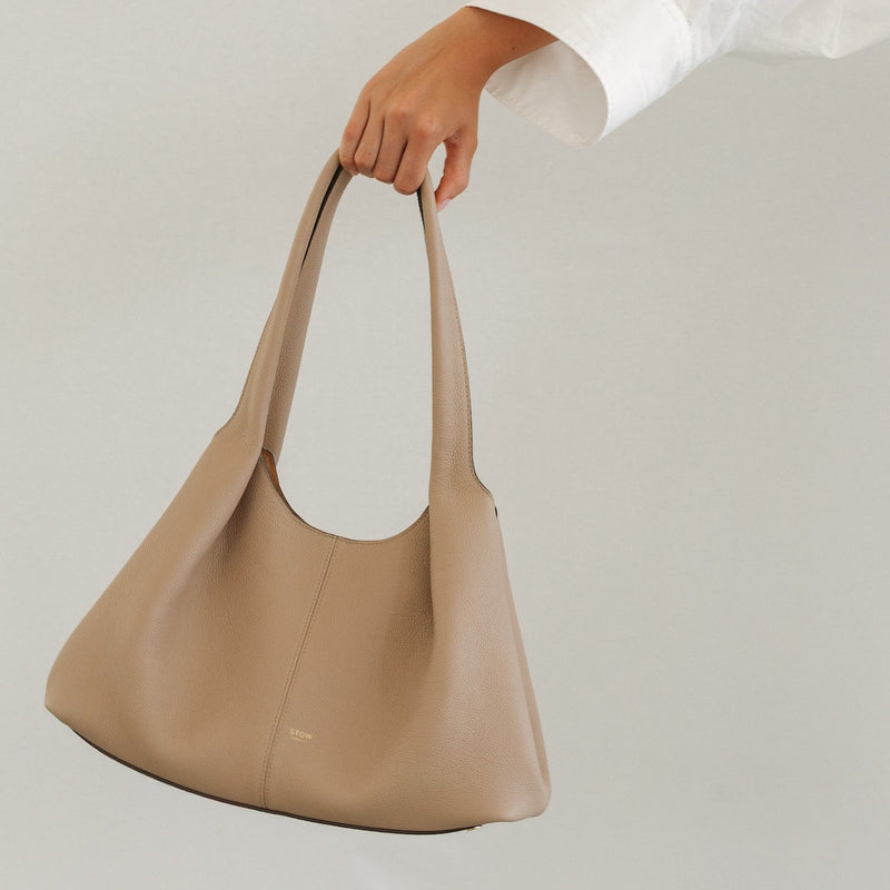 Model holding STOW London's Hobo Tote in Camel pebbled leather featuring a pleated front panel and gold-coated stainless steel hardware 