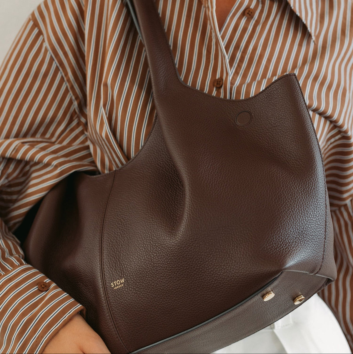 Model holding STOW London's Hobo Tote in Espresso pebbled leather featuring a pleated front panel and gold-coated stainless steel hardware 