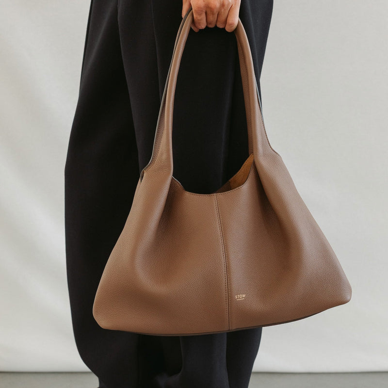 Model holding STOW London's Hobo Tote in Mocha pebbled leather featuring a pleated front panel and gold-coated stainless steel hardware 