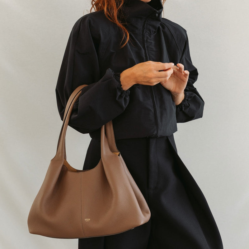 Model holding STOW London's Hobo Tote in Mocha pebbled leather featuring a pleated front panel and gold-coated stainless steel hardware 