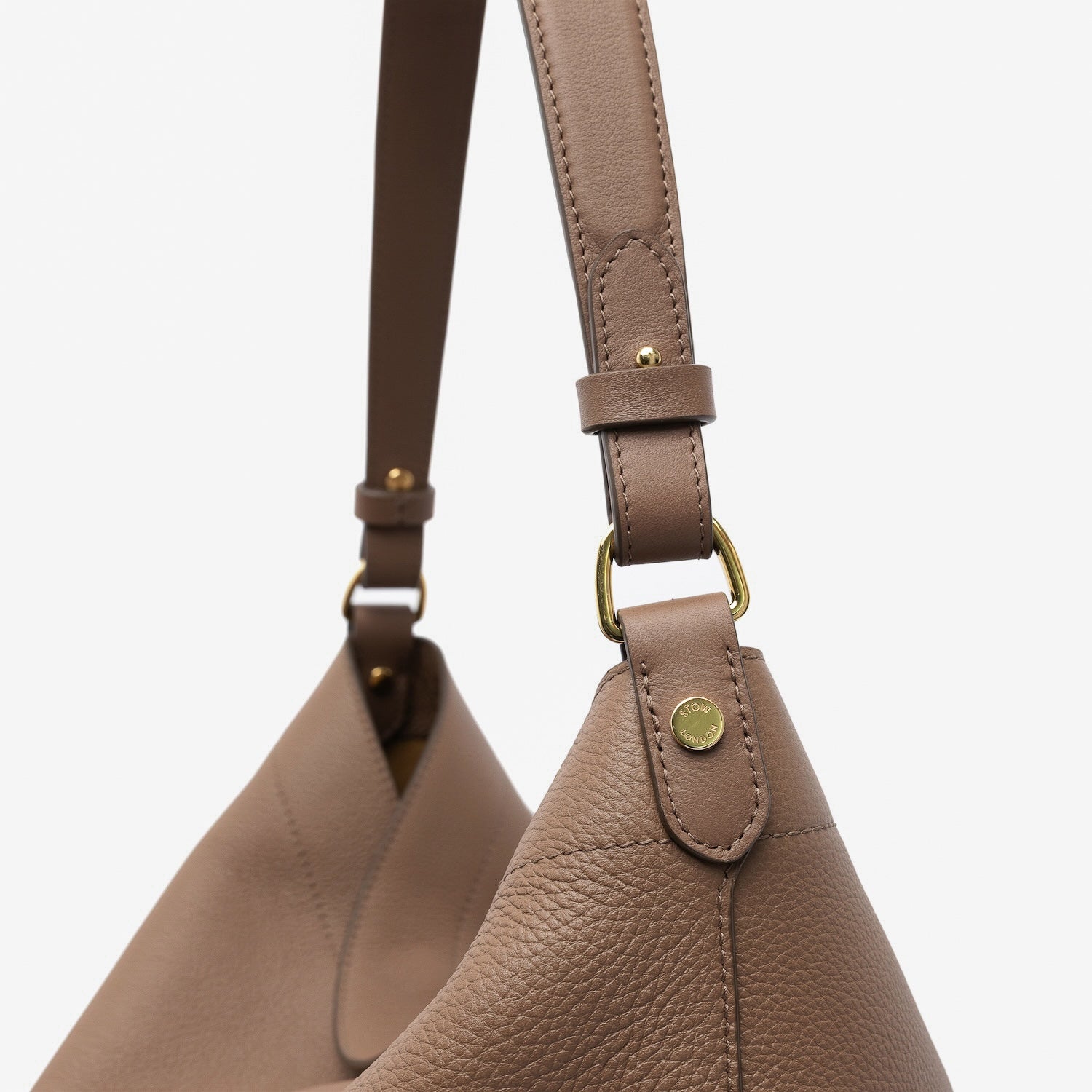 Close up of the handles on STOW London's Maxi Curve bag in Mocha pebbled leather featuring gold-coated stainless steel hardware on a light grey background