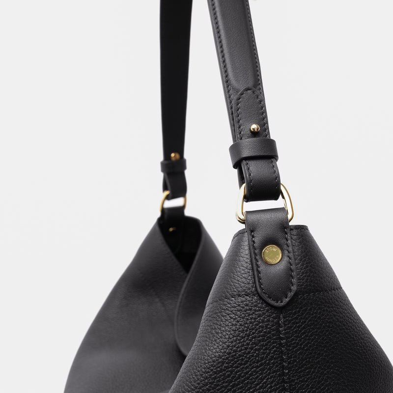 Close up of STOW London's Maxi Curve bag in Black pebbled leather featuring gold-coated stainless steel hardware on a light grey background