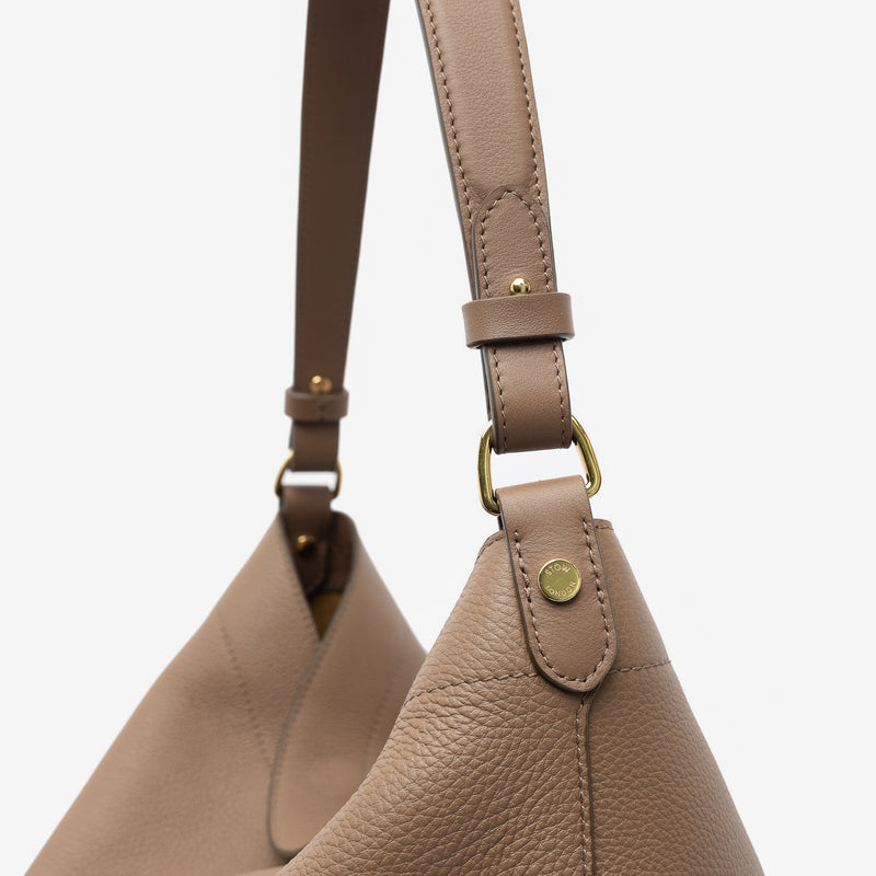 Close up of the handles on STOW London's Maxi Curve bag in Mocha pebbled leather featuring gold-coated stainless steel hardware on a light grey background