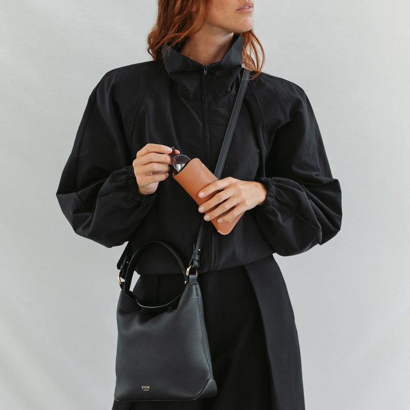 Model wearing STOW London's Midi Curve bag in Black pebbled and smooth leather crossbody featuring gold-coated stainless steel hardware and the Earth Tan Glasses Case