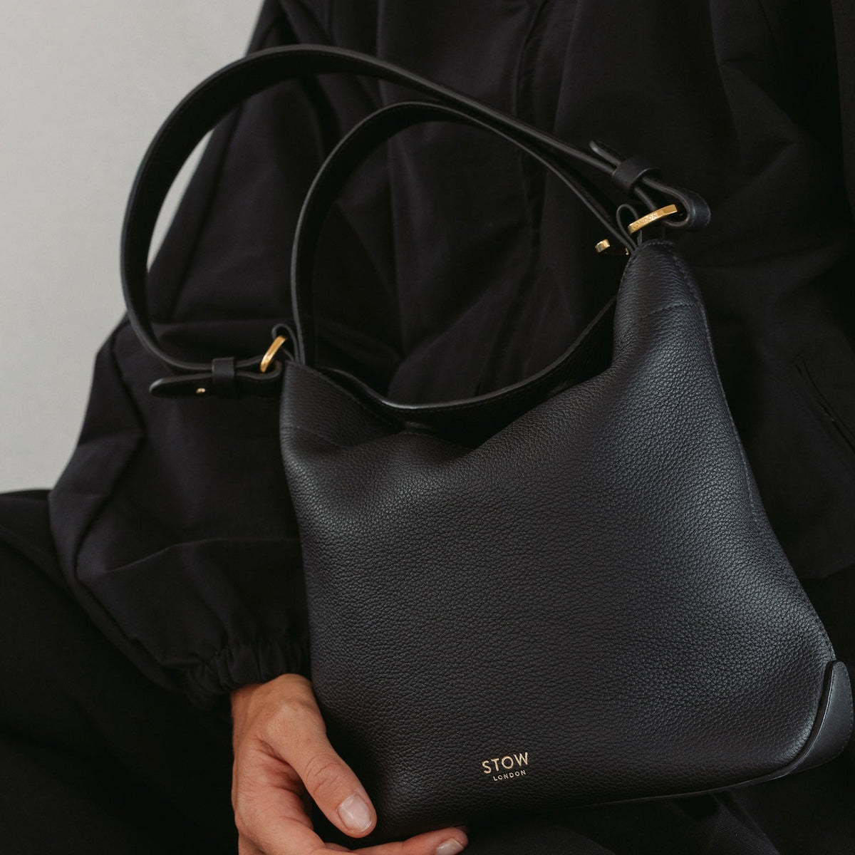 STOW London's Midi Curve in Black pebbled leather with gold hardware and a small gold logo, held by a model wearing a black jacket