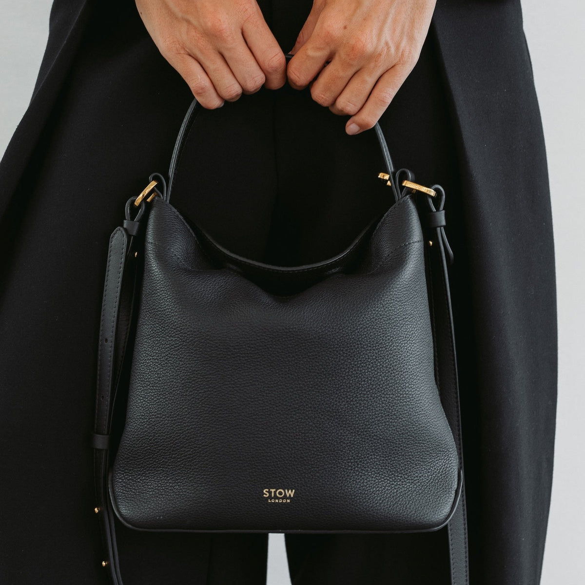 Model holding STOW London's Midi Curve bag in Black pebbled and smooth leather at her front in a classic flap bag style featuring gold-coated stainless steel hardware