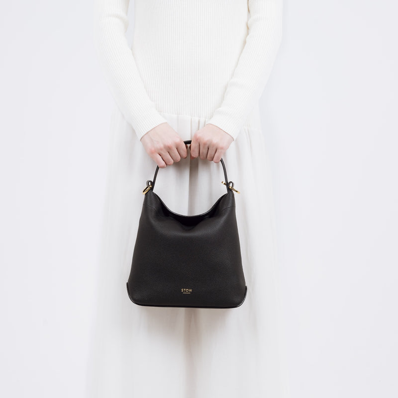 Model holding STOW London's Midi Curve bag in Black pebbled and smooth leather at her front in a classic flap bag style featuring gold-coated stainless steel hardware on a white background