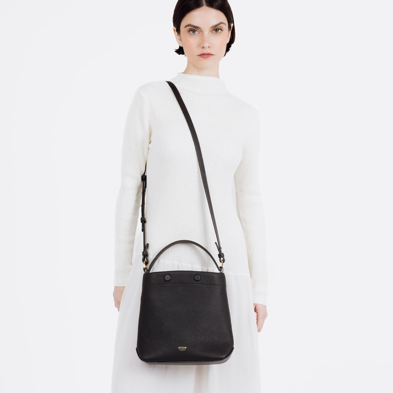 Model wearing STOW London's Midi Curve bag in Black pebbled and smooth leather over her shoulder in a bucket bag style featuring gold-coated stainless steel hardware on a white background