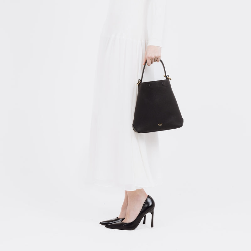 Model holding STOW London's Midi Curve bag in Black pebbled and smooth leather by the top handle at her side styled as a bucket bag featuring gold-coated stainless steel hardware on a white background