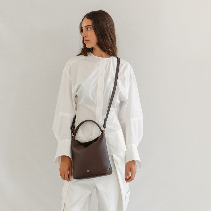 Model in a white outfit wearing STOW London's Midi Curve in Espresso pebbled leather crossbody featuring gold-coated stainless steel hardware 