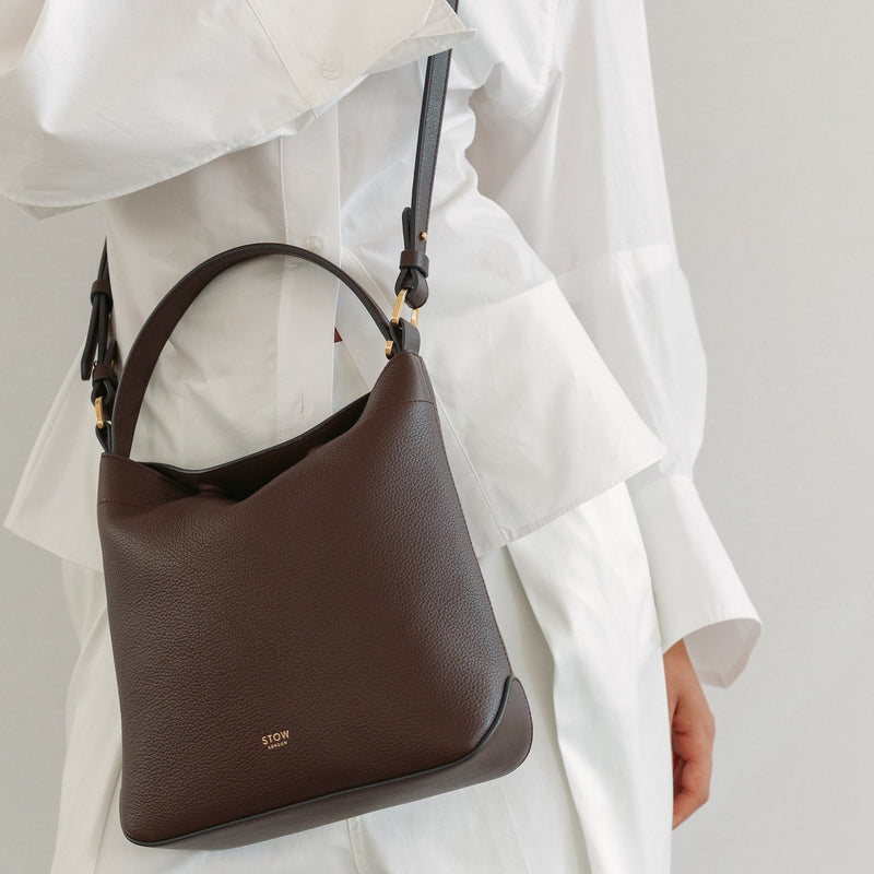 Model in a white outfit wearing STOW London's Midi Curve in Espresso pebbled leather crossbody featuring gold-coated stainless steel hardware 