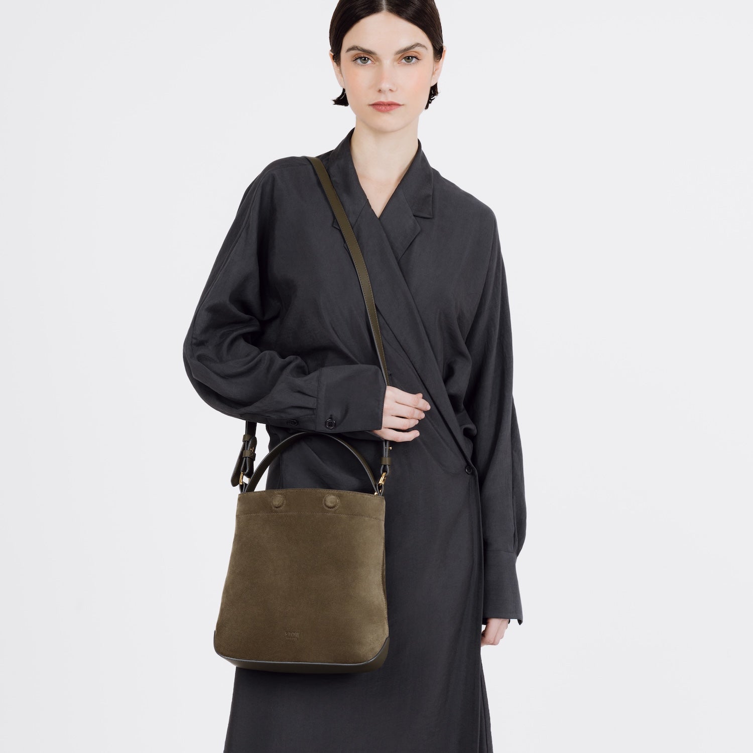 Model wearing STOW London's Midi Curve bag in Dark Green Suede leather on her shoulder styled as a bucket bag featuring gold-coated stainless steel hardware on a white background