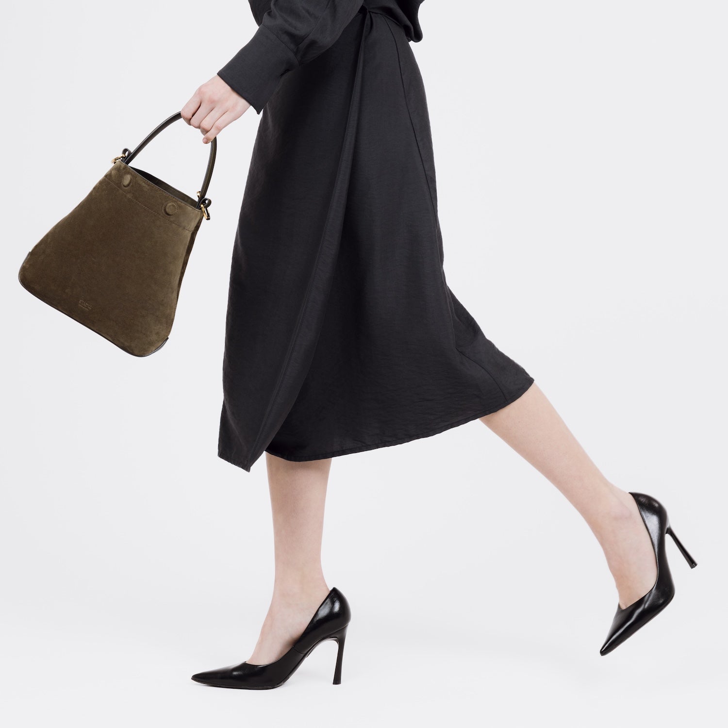 Model walking with STOW London's Midi Curve bag in Dark Green Suede leather in her hand styled as a bucket bag featuring gold-coated stainless steel hardware on a white background