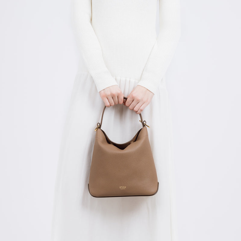 Model holding STOW London's Midi Curve bag in Mocha pebbled and smooth leather by the top handle at her front as a flap bag featuring gold-coated stainless steel hardware on a white background