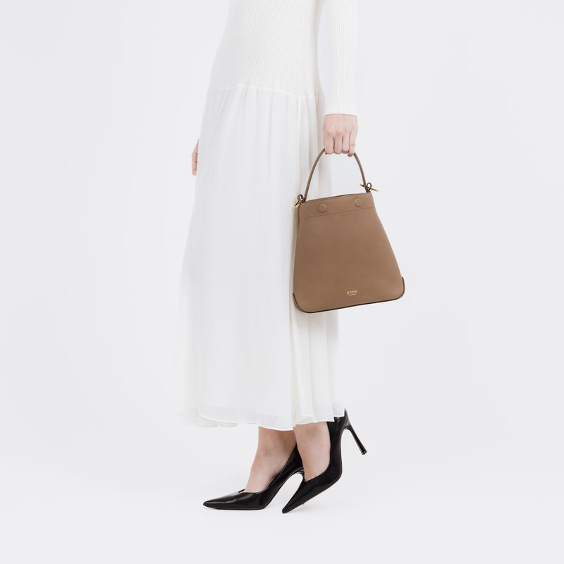 Model holding STOW London's Midi Curve bag in Mocha pebbled and smooth leather by the top handle at her side featuring gold-coated stainless steel hardware on a white background