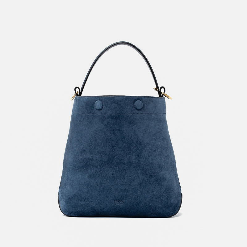 STOW London's Midi Curve in Navy suede leather styled as a bucket bag featuring gold-coated stainless steel hardware on a light grey background