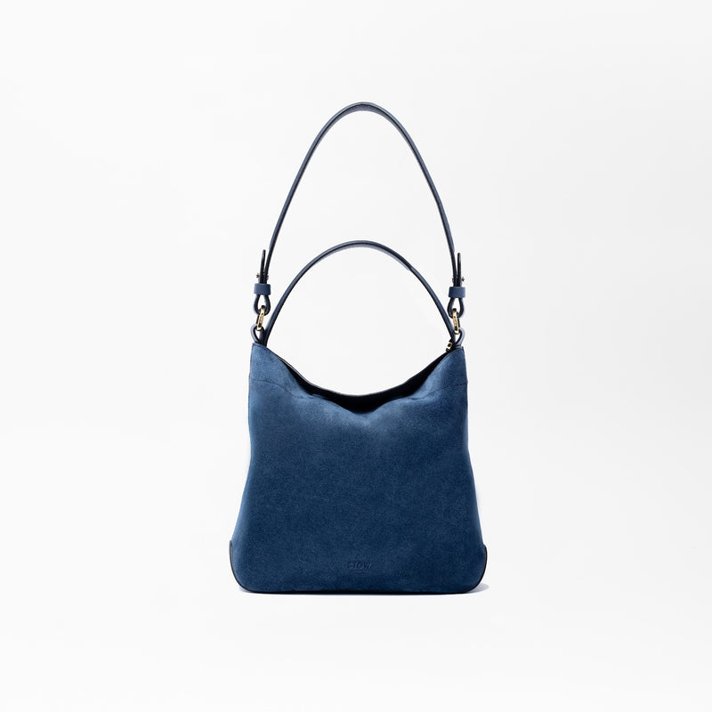 STOW London's Midi Curve in Navy suede leather on a light grey background featuring the detachable shoulder strap and top handle with gold hardware and details 
