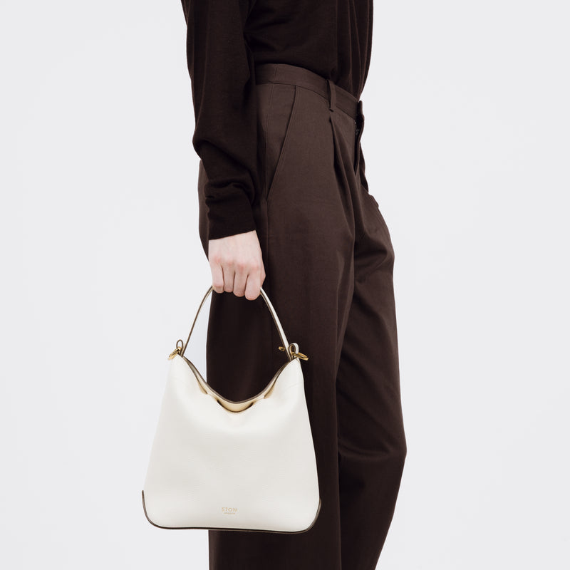 Model holding STOW London's Midi Curve bag in Spring Moon pebbled and smooth leather at her side styled as a flap bag featuring gold-coated stainless steel hardware on a white background