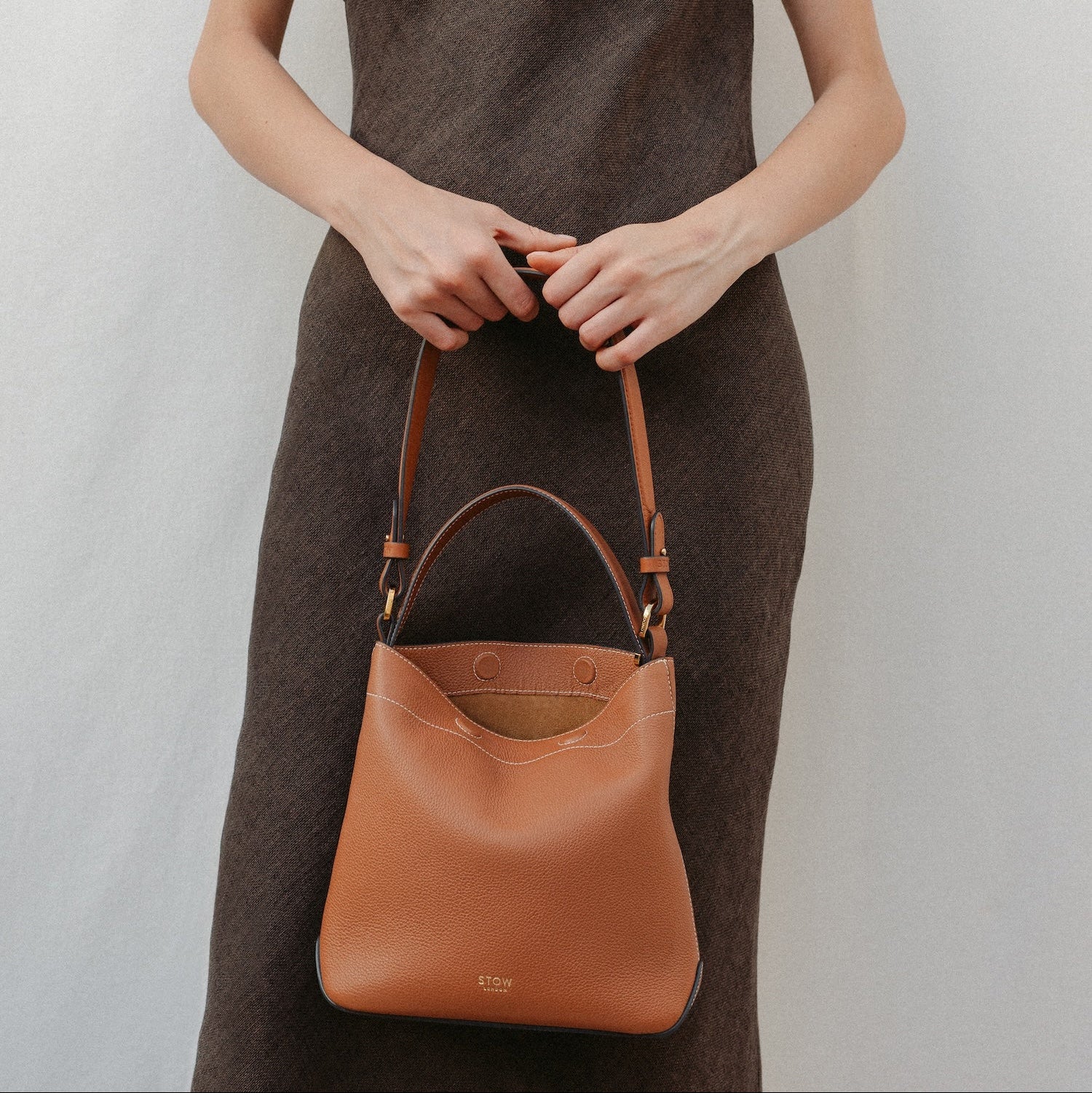 Model holding STOW London's Curve in Earth Tan leather featuring the detachable shoulder strap and top handle with gold hardware and details 