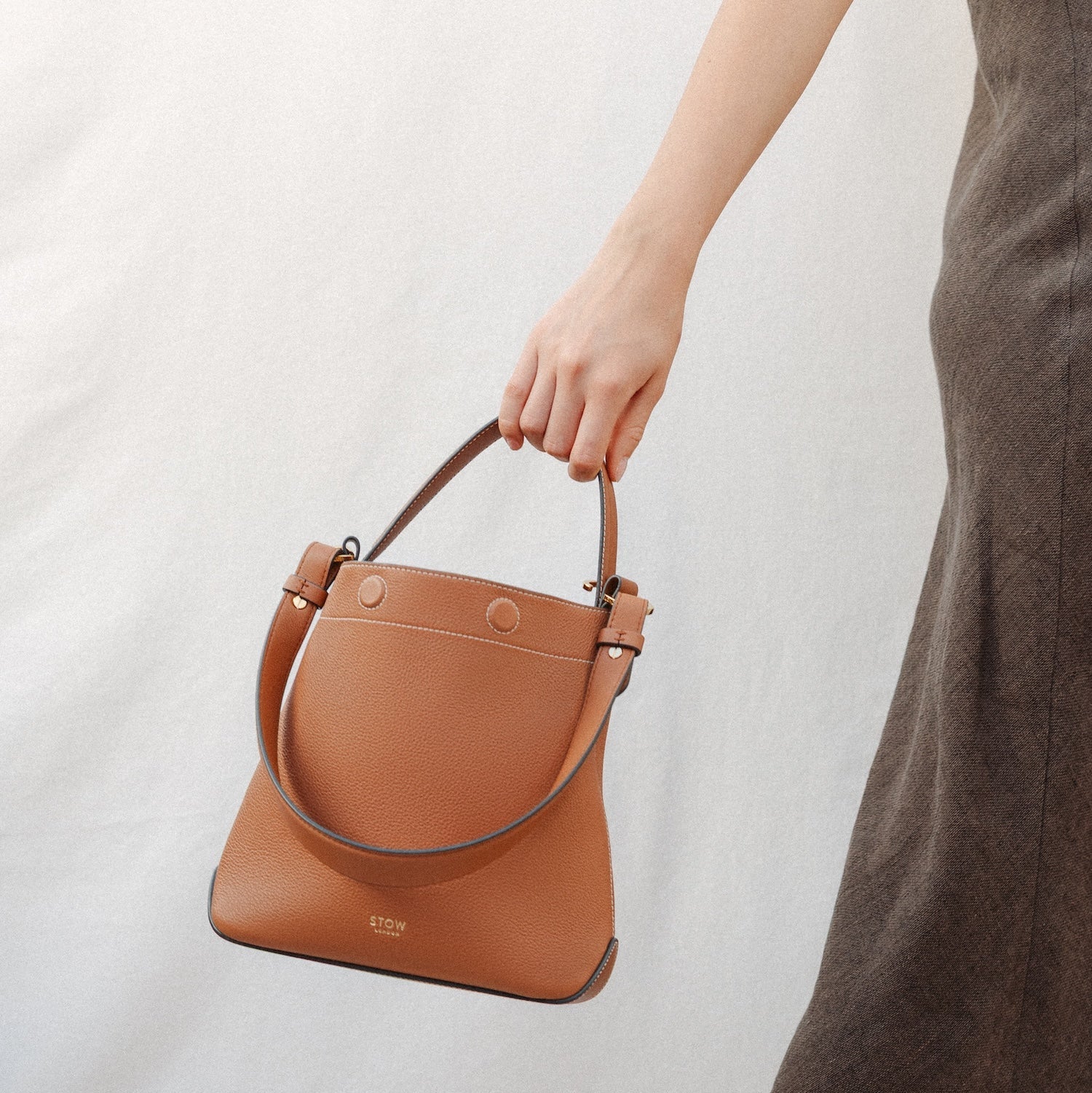 Model holding STOW London's Curve in Earth Tan leather featuring the detachable shoulder strap with gold hardware and details 