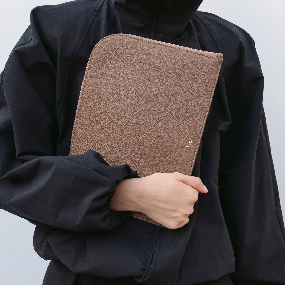 Model holding STOW London's Midi Leather Sleeve in Mocha pebbled leather featuring a small gold embossed logo on the front 