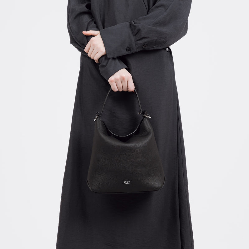 Model holding STOW London's Midi Curve bag in Black pebbled and smooth leather by the top handle at her front in the flap bag style featuring silver stainless steel hardware on a white background