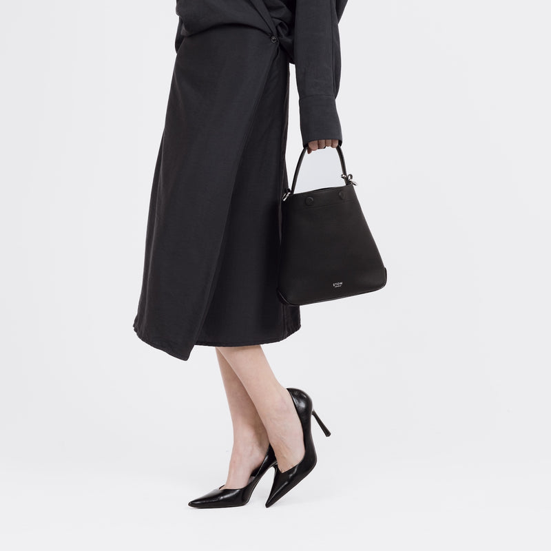 Model wearing STOW London's Midi Curve bag in Black pebbled and smooth leather by the top handle at her side in a bucket bag style featuring silver stainless steel hardware on a white background