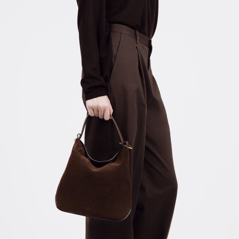 Model holding STOW London's Midi Curve bag in Chocolate Suede leather at her side shaped as a flap bag featuring gold-coated stainless steel hardware on a white background