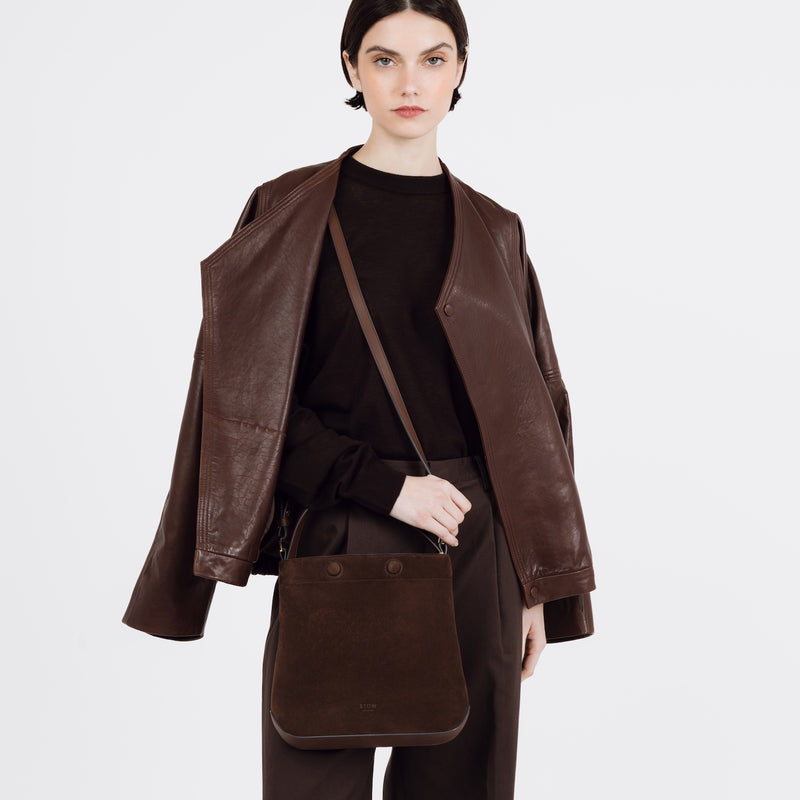 Model wearing STOW London's Midi Curve bag in Chocolate Suede leather on her shoulder styled as a bucket bag featuring gold-coated stainless steel hardware on a white background
