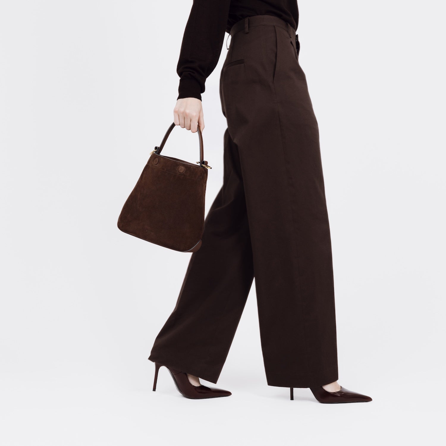 Model holding STOW London's Midi Curve bag in Chocolate Suede leather at her side styled as a bucket bag featuring gold-coated stainless steel hardware on a white background