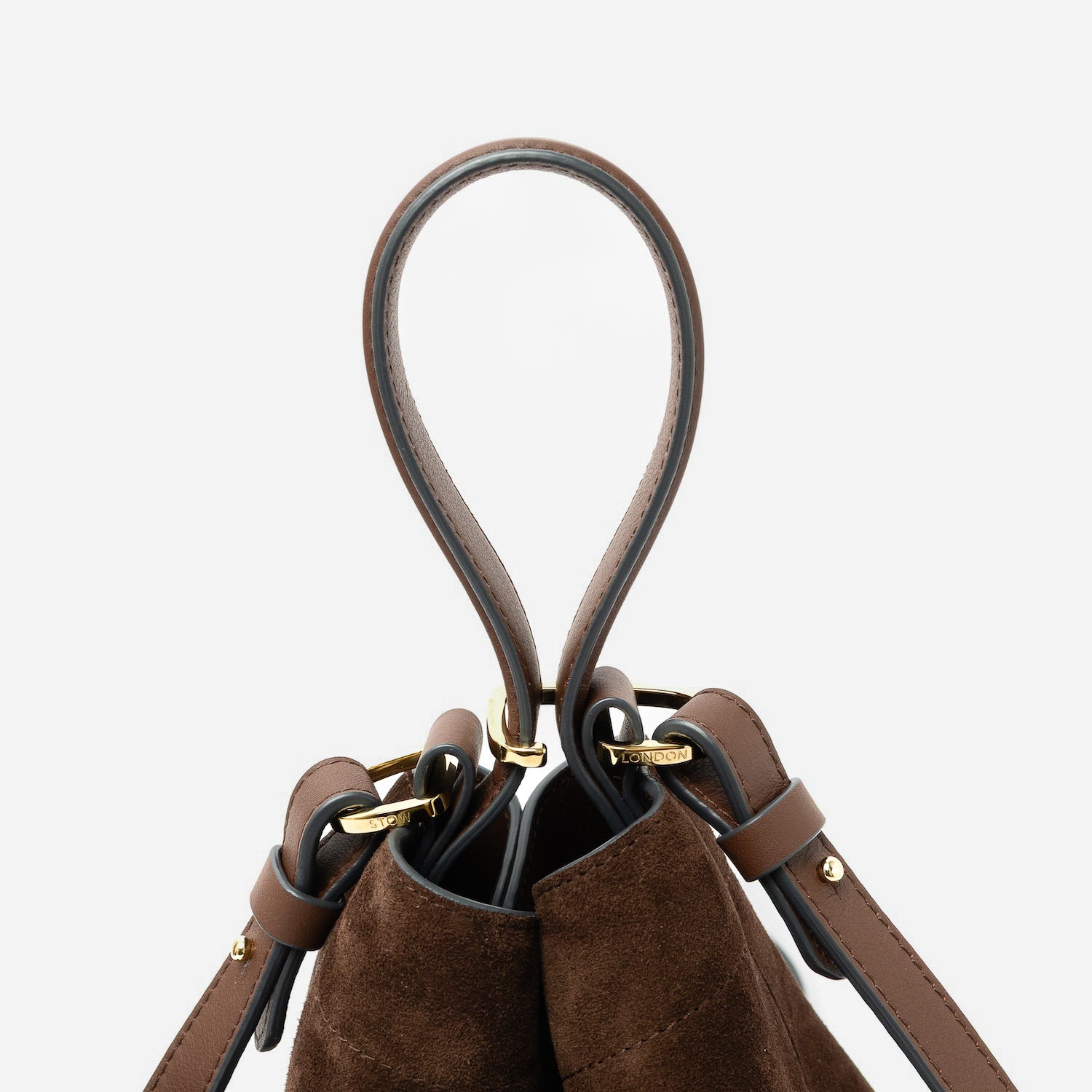 Close up of the handles of STOW London's Midi Curve in Chocolate suede leather featuring gold-coated stainless steel hardware, clip and detachable strap on a light grey background