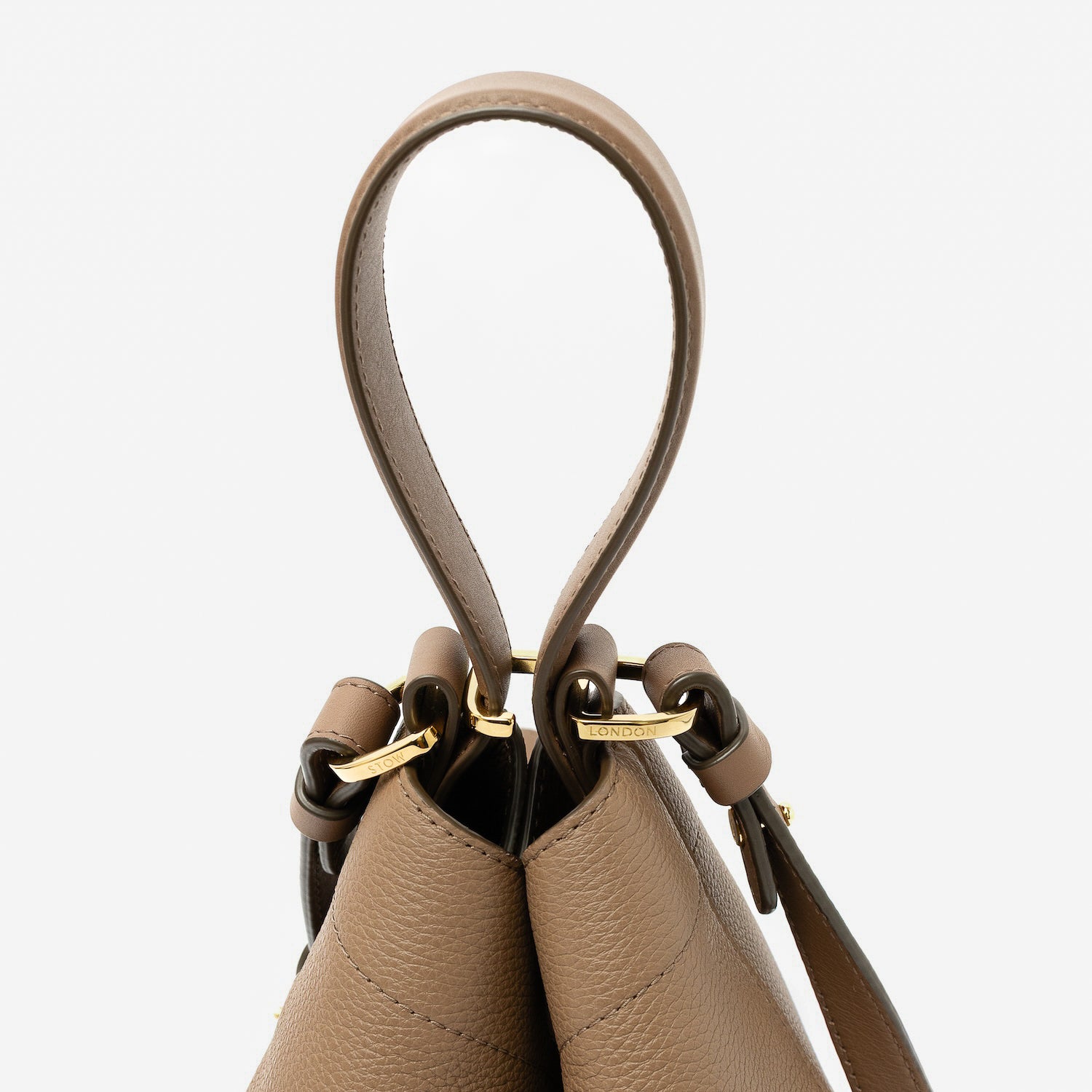 Close up of the handles on STOW London's Midi Curve in Mocha pebbled leather featuring gold-coated stainless steel hardware, clip and detachable strap on a light grey background