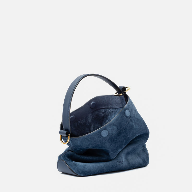 Off-axis view of STOW London's Midi Curve in Navy suede leather with magnets open showing suede interior featuring gold-coated stainless steel hardware on a light grey background