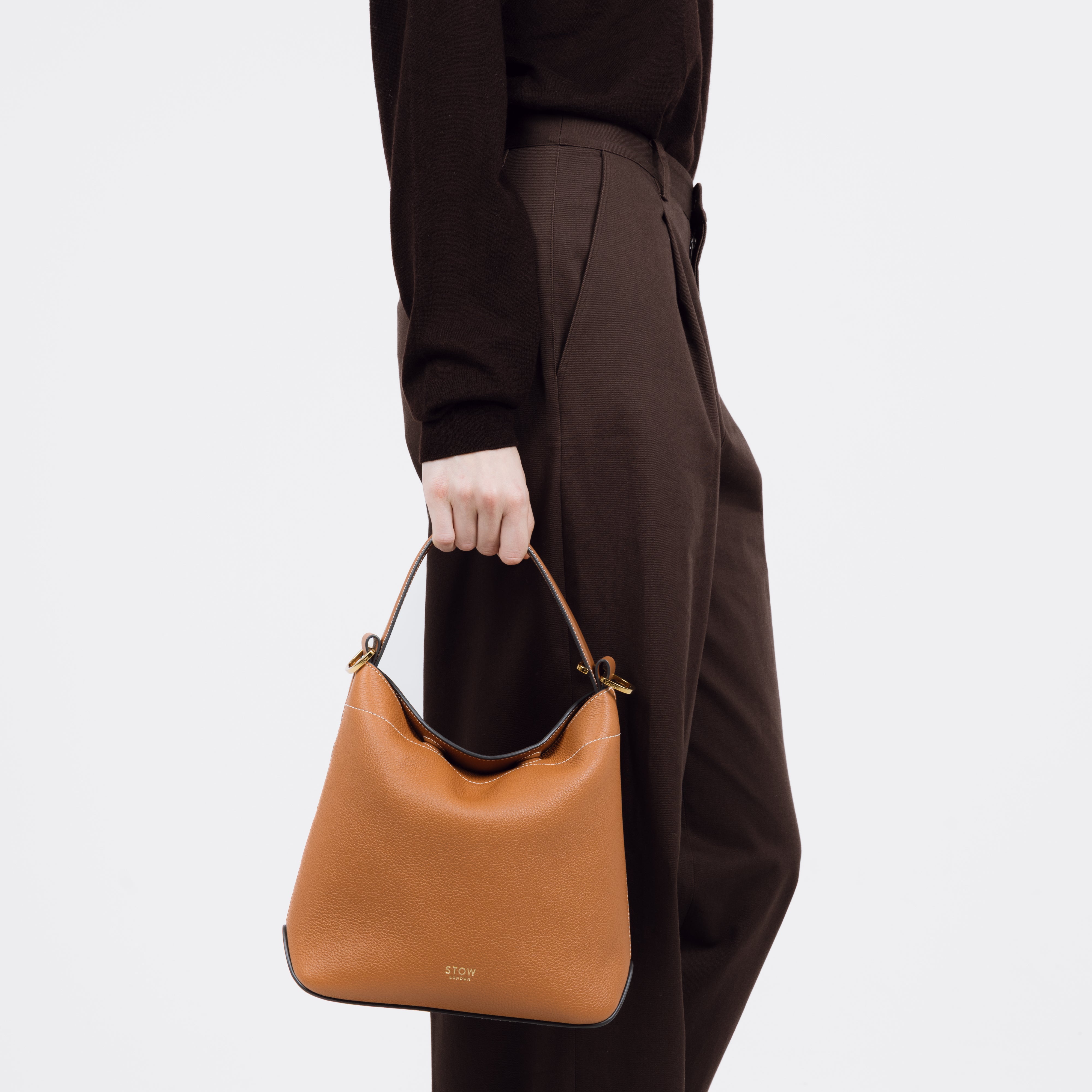 Model holding STOW London's Midi Curve bag in Earth Tan pebbled and smooth leather at her side shaped as a flap bag featuring gold-coated stainless steel hardware on a white background