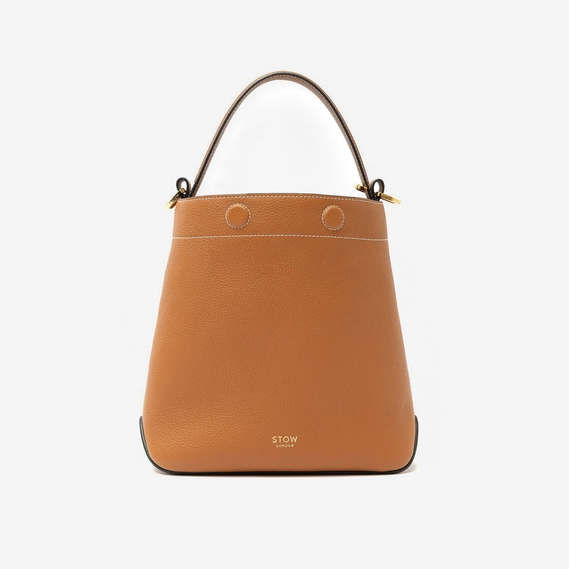 STOW London's Midi Curve in Earth Tan pebbled leather styled as a bucket bag featuring gold-coated stainless steel hardware on a light grey background